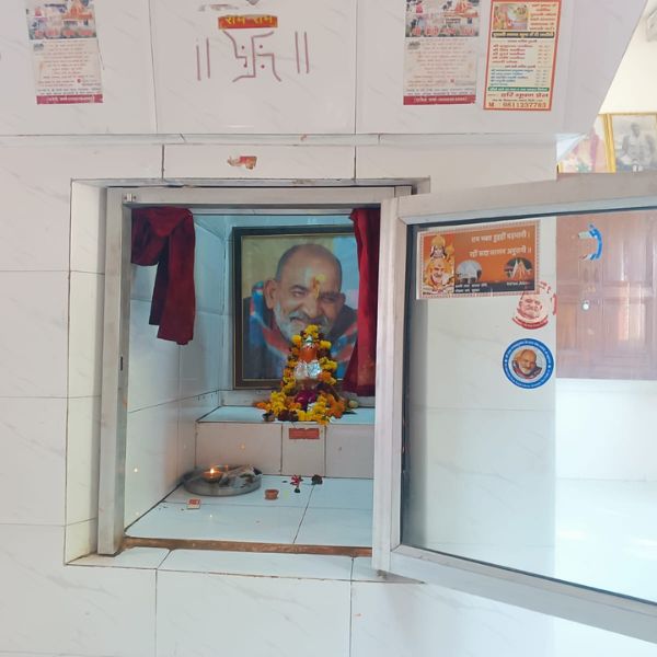 Delhi ke Neem Karoli Baba mandir me Maharaj ji ki muskurati hui tasveer, jiske aage shraddha se phool aur diya rakha hua hai.